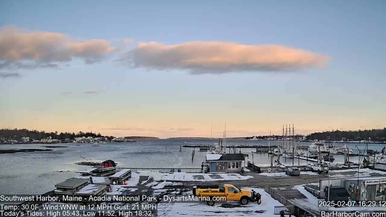 A snow-covered harbor with boats docked, a yellow truck, and a partly cloudy sunset.