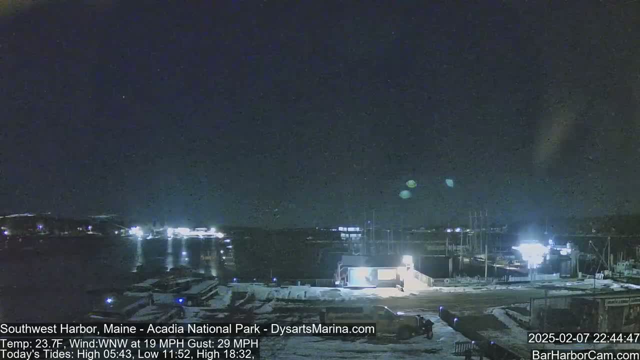 A nighttime view of a harbor with boats and a building, under a dark sky with a few faint lights visible, and snow on the ground.