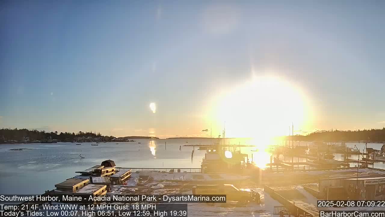 A sunrise over a harbor with boats moored, partially covered in snow, under a mostly clear sky and a wind of 12 mph gusting to 18 mph.
