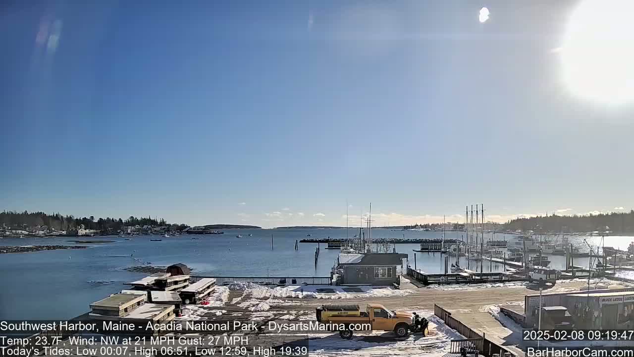 A sunny harbor scene with snow on the ground and a yellow snowplow, boats in the water, and a clear blue sky.