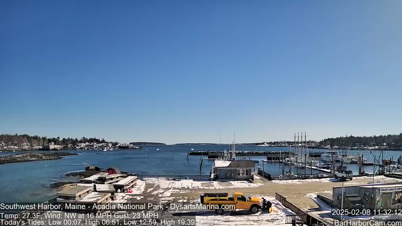 A snow-dusted harbor scene shows numerous boats docked at a marina, under a clear blue sky.