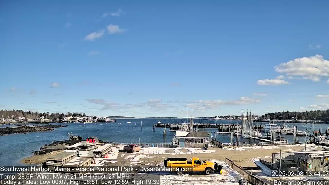 A harbor scene with a partly sunny sky, a light covering of snow on the ground, and a yellow truck in a parking lot next to docked sailboats and buildings.