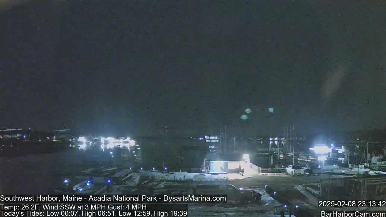 A nighttime view of a harbor with boats docked, under a dark sky with a few faint lights visible in the distance, and a light snow cover on the ground.