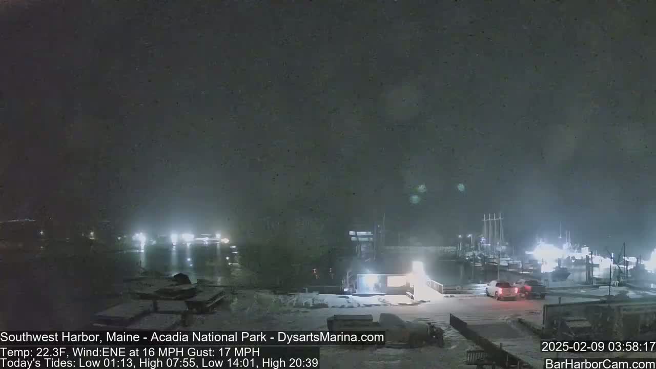 A snow-covered harbor at night, with boats docked and a few vehicles parked nearby under a dark, cloudy sky.