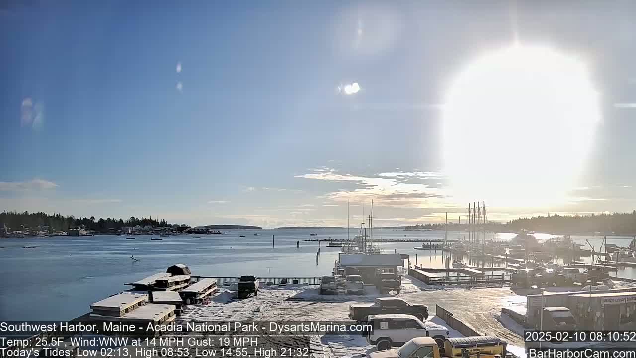 A sunny, cold day with snow on the ground shows a harbor filled with boats and cars parked in a lot next to the water.