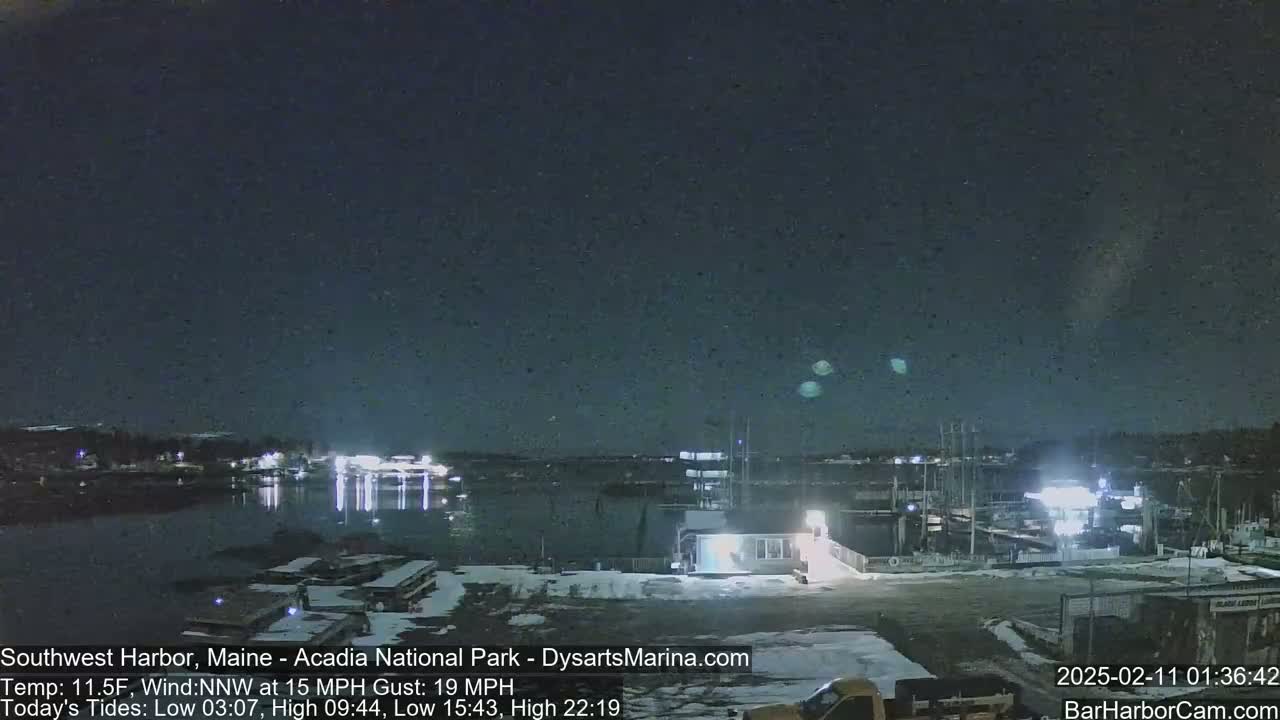 A nighttime view of a snow-covered harbor with a calm body of water reflecting lights from buildings and boats under a clear, dark sky.