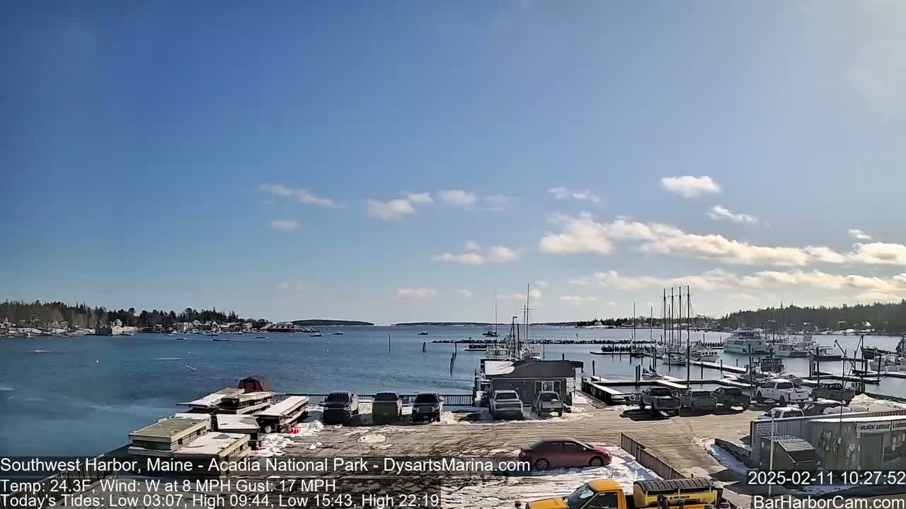 A harbor with numerous boats and cars parked in a snow-dusted parking lot is visible under a mostly sunny sky with some scattered clouds.