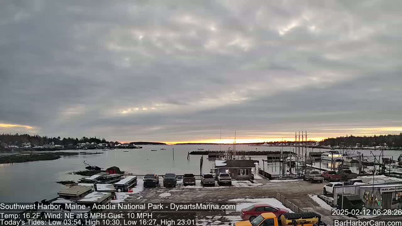 A partly cloudy sunrise over a harbor with boats docked and cars parked in a snow-covered lot.