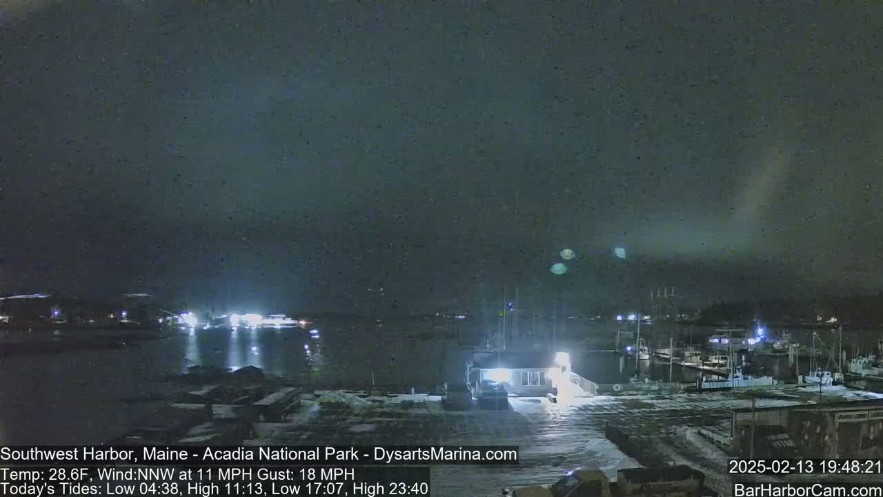 A nighttime harbor scene with snow on the ground and a dark, cloudy sky, under which boats are docked near lit buildings.