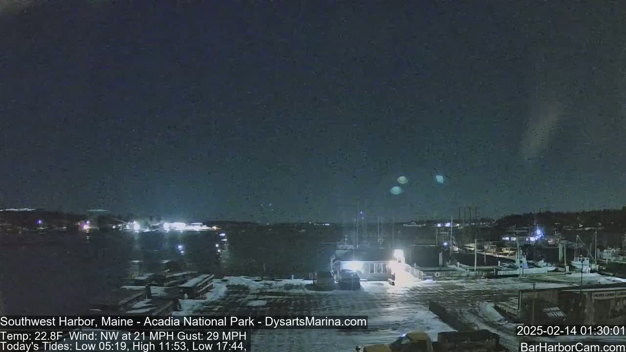 A nighttime view of a snow-covered harbor with boats and buildings under a dark sky with a few faint lights visible.