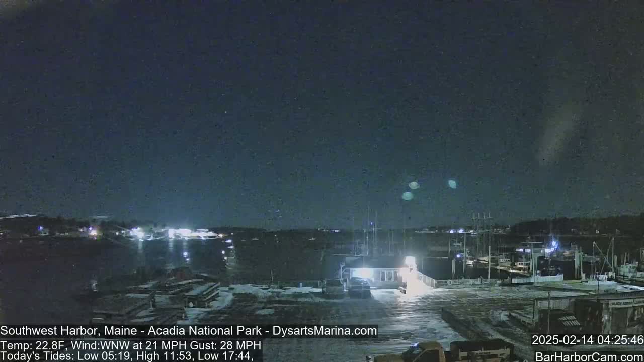 A nighttime harbor scene with snow on the ground, under a dark sky with three bright lights visible in the distance, and a windy, cold temperature.