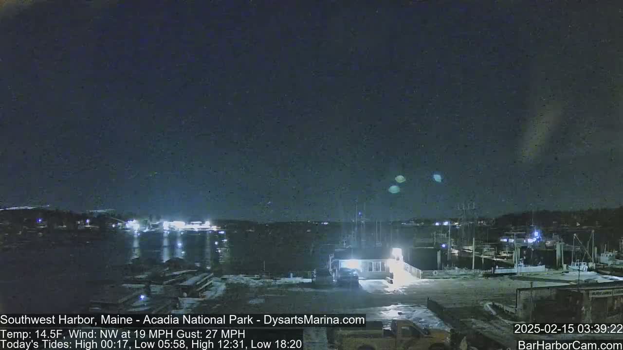 A nighttime view of a harbor with boats, buildings, and vehicles under a dark sky with a few faint lights, and a northwest wind at 19 mph with gusts up to 27 mph.