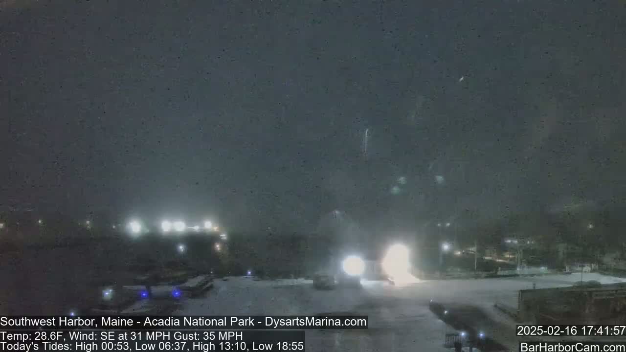 A nighttime snowstorm with strong southeast winds illuminates a snow-covered parking lot and buildings with bright lights.