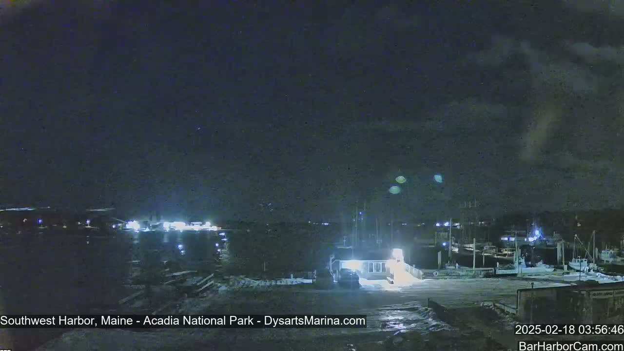 A nighttime view of a harbor with boats docked, illuminated by lights, under a mostly cloudy sky.