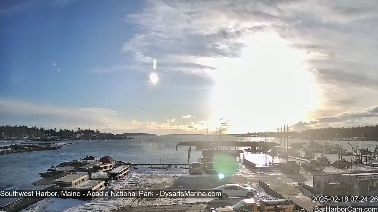 A partly sunny day shows a harbor with boats and a town in the background, snow on the ground near the foreground.