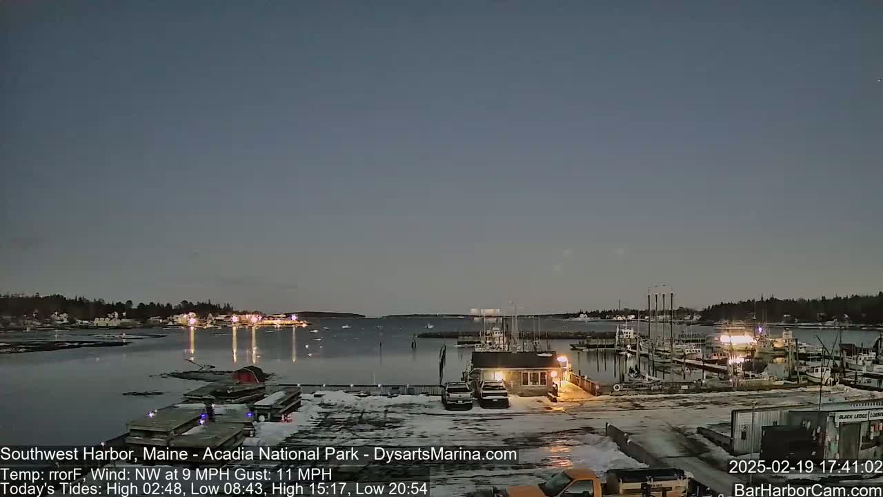 A twilight view of a harbor with boats docked, some snow on the ground, and a calm, dark sky.