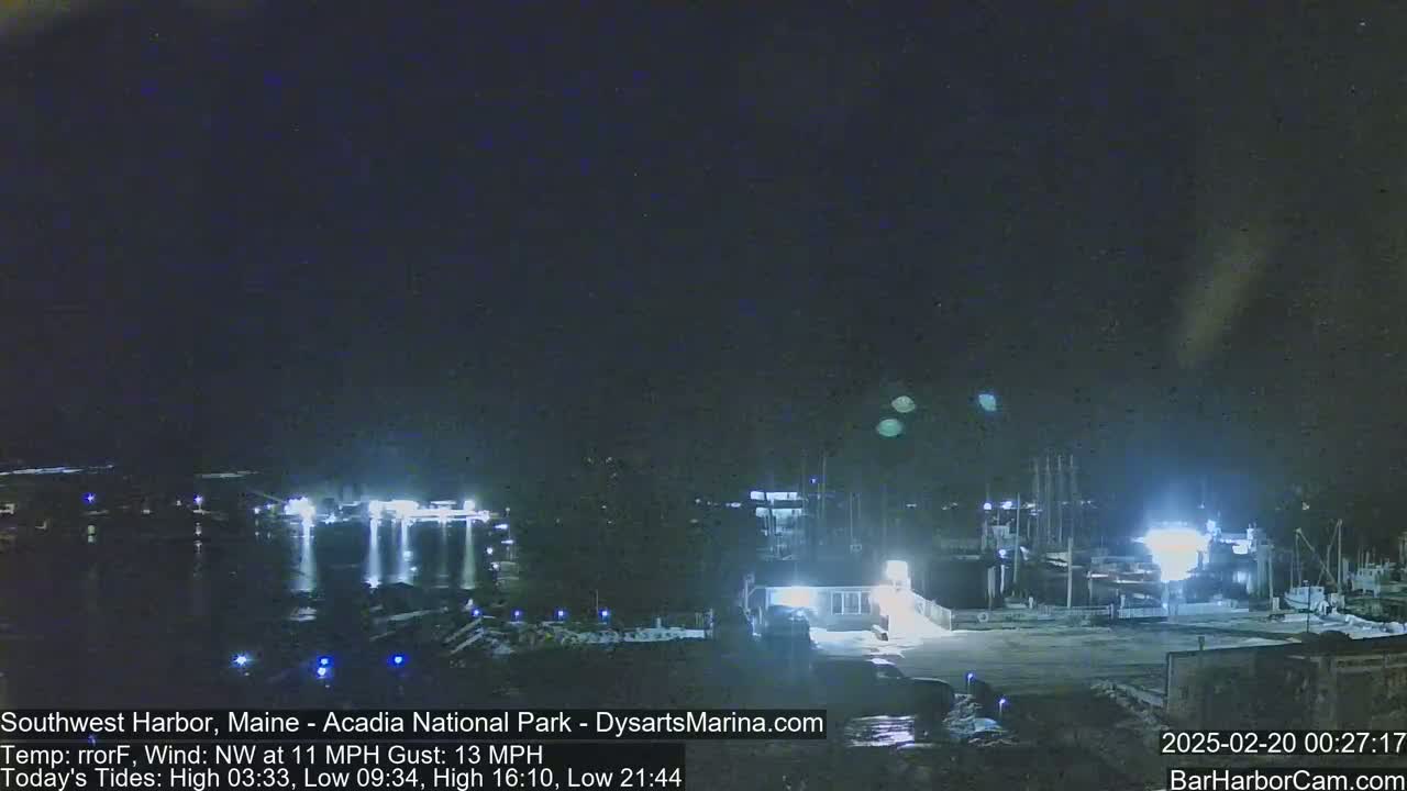 A nighttime harbor scene with boats docked, illuminated by lights, under a dark sky with a northwest wind.