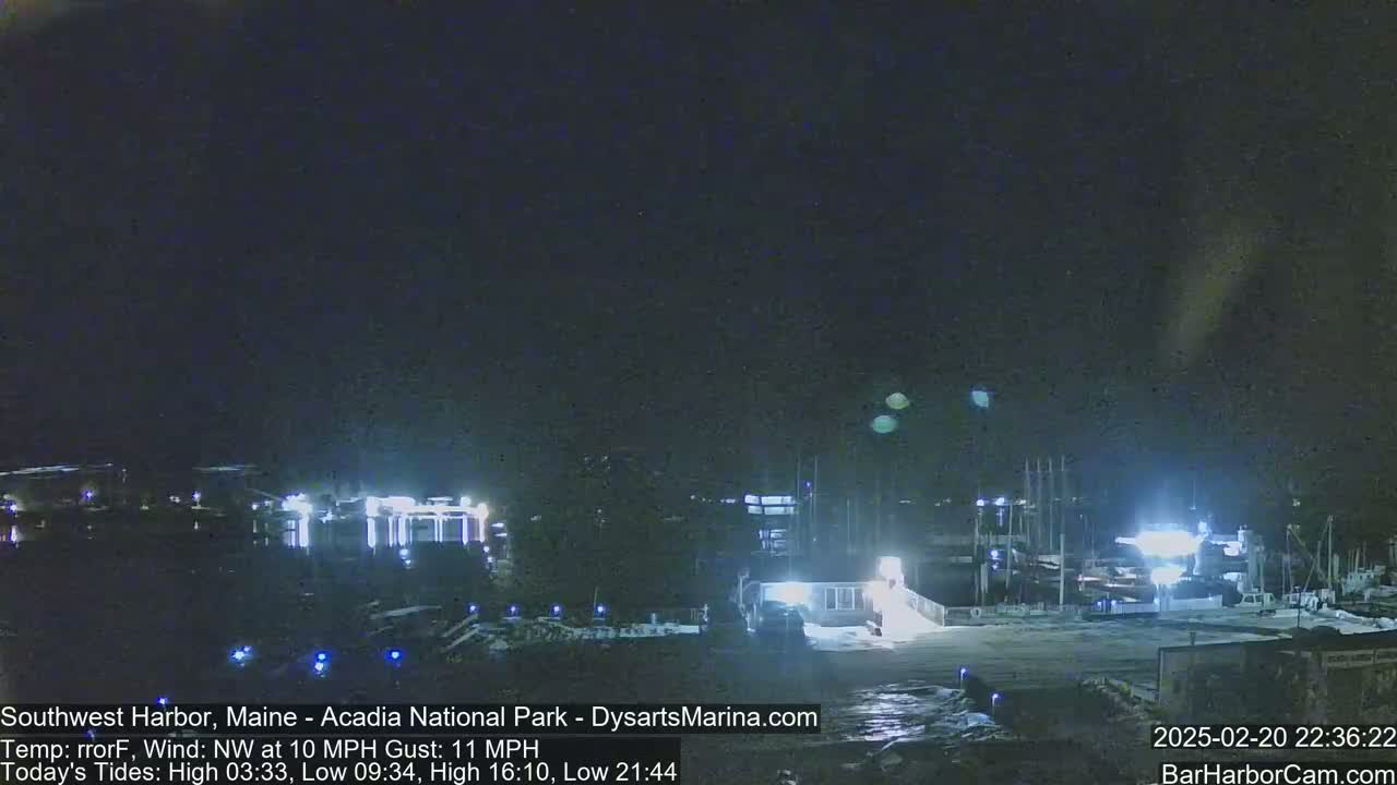 A nighttime harbor scene with a northwest wind at 10 mph gusting to 11 mph, showing boats docked and buildings lit up.