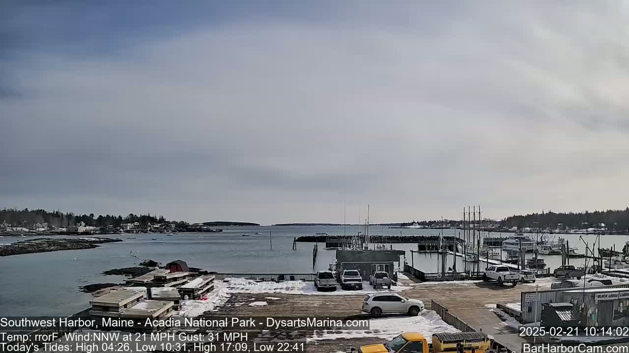 A partly sunny day shows a harbor with docked boats and cars parked in a lot near the water's edge, with a town visible across the water.