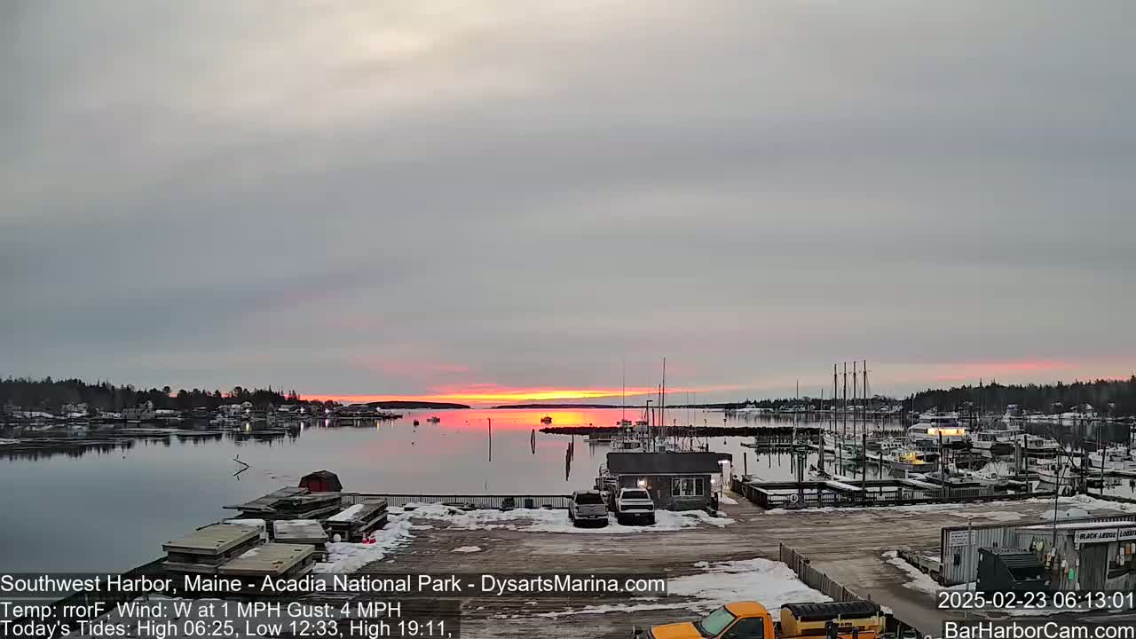 A calm harbor with docked boats is seen at sunset under a mostly cloudy sky, with some snow visible on the ground.