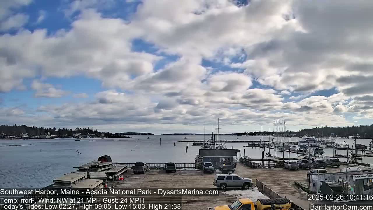 A partly cloudy day shows a harbor with numerous boats docked, some cars parked on a nearby wooden pier, and a town across the water.
