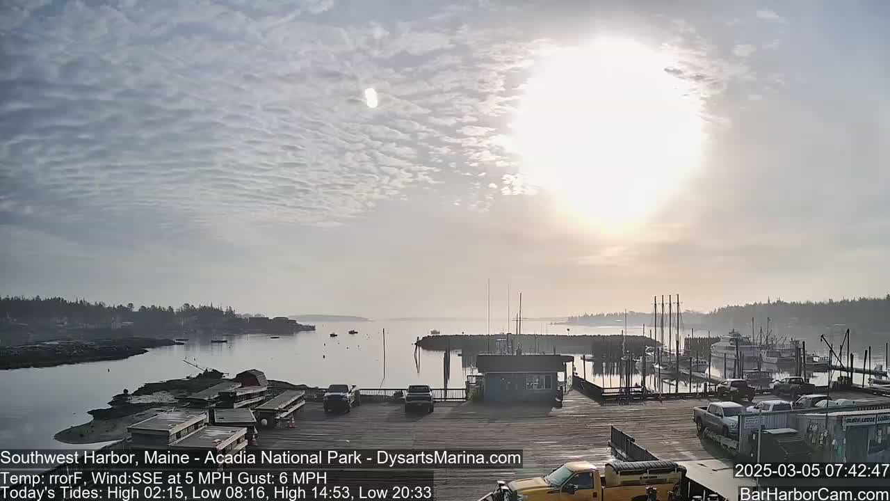 A harbor with boats and a dock is visible under a mostly cloudy sky with the sun shining brightly through the clouds.