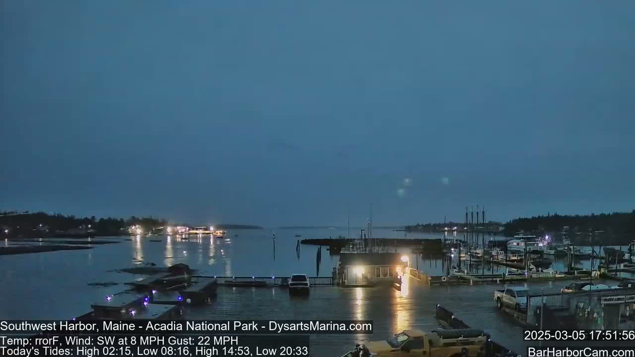 A dark, wet harbor at twilight with boats moored at a marina and a few vehicles parked nearby, under a mostly clear sky with a southwest wind.