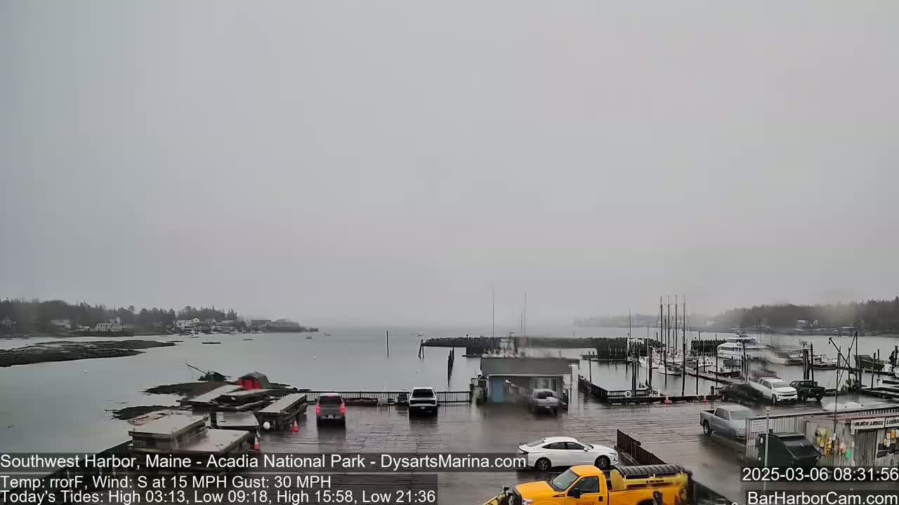 A mostly cloudy and wet day at a harbor with boats docked and several cars parked in a lot.