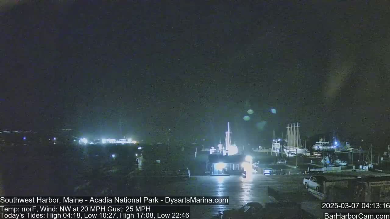 A nighttime harbor scene with boats docked, a building lit up, and a vehicle parked nearby, under a dark, slightly cloudy sky with a northwest wind.