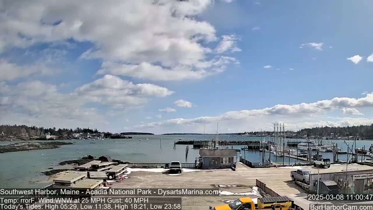 A partly cloudy day shows a harbor with many boats docked, some vehicles in a parking lot, and a small amount of snow on the ground.