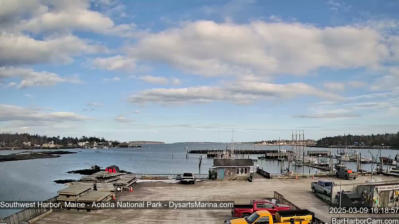 A partly cloudy day reveals a harbor filled with boats, a few cars in a parking lot, and a small town across the water.