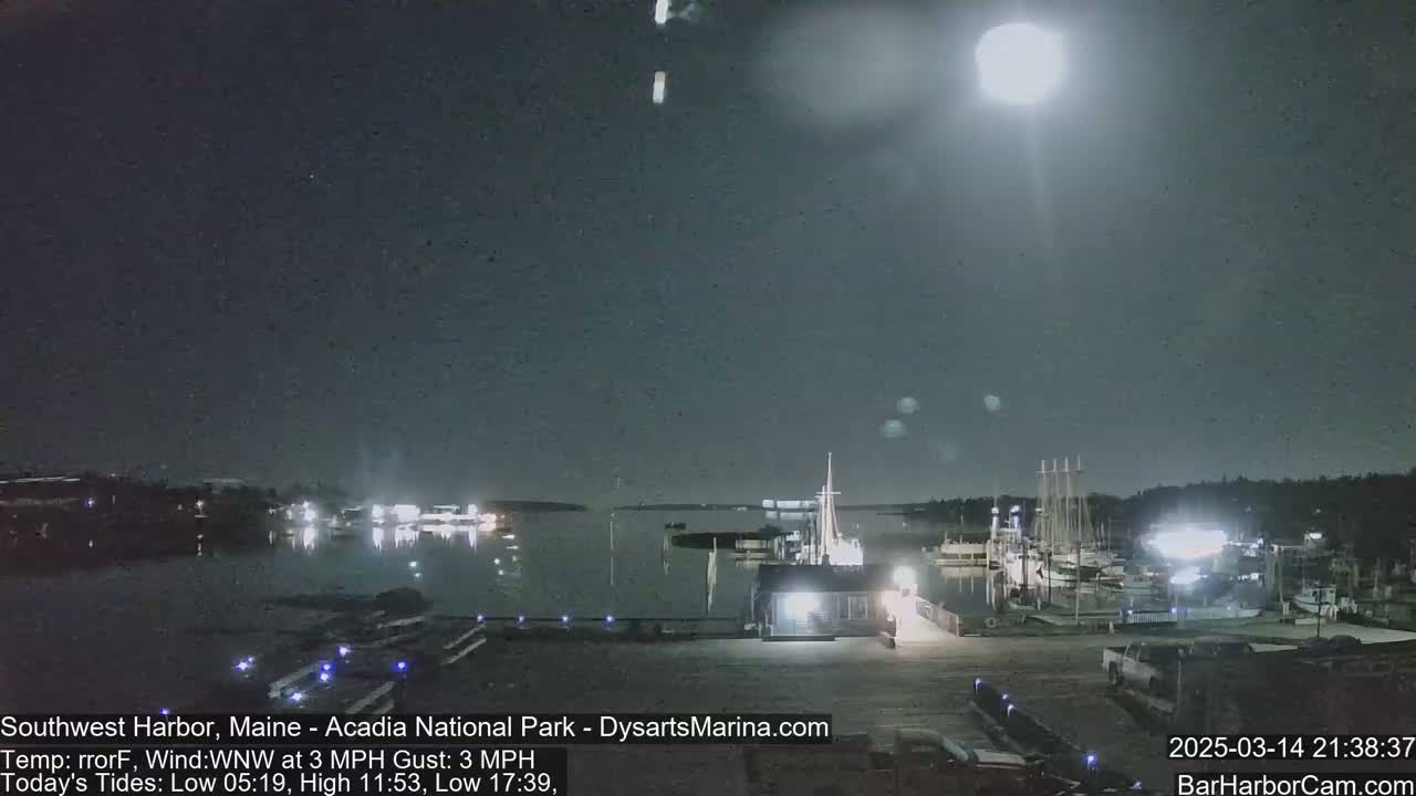 A nighttime view of a harbor with boats docked, under a mostly clear sky illuminated by a bright light source.