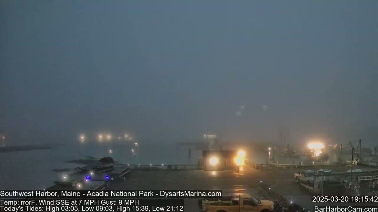 A foggy harbor scene at night shows lights illuminating a dock with several vehicles and boats.