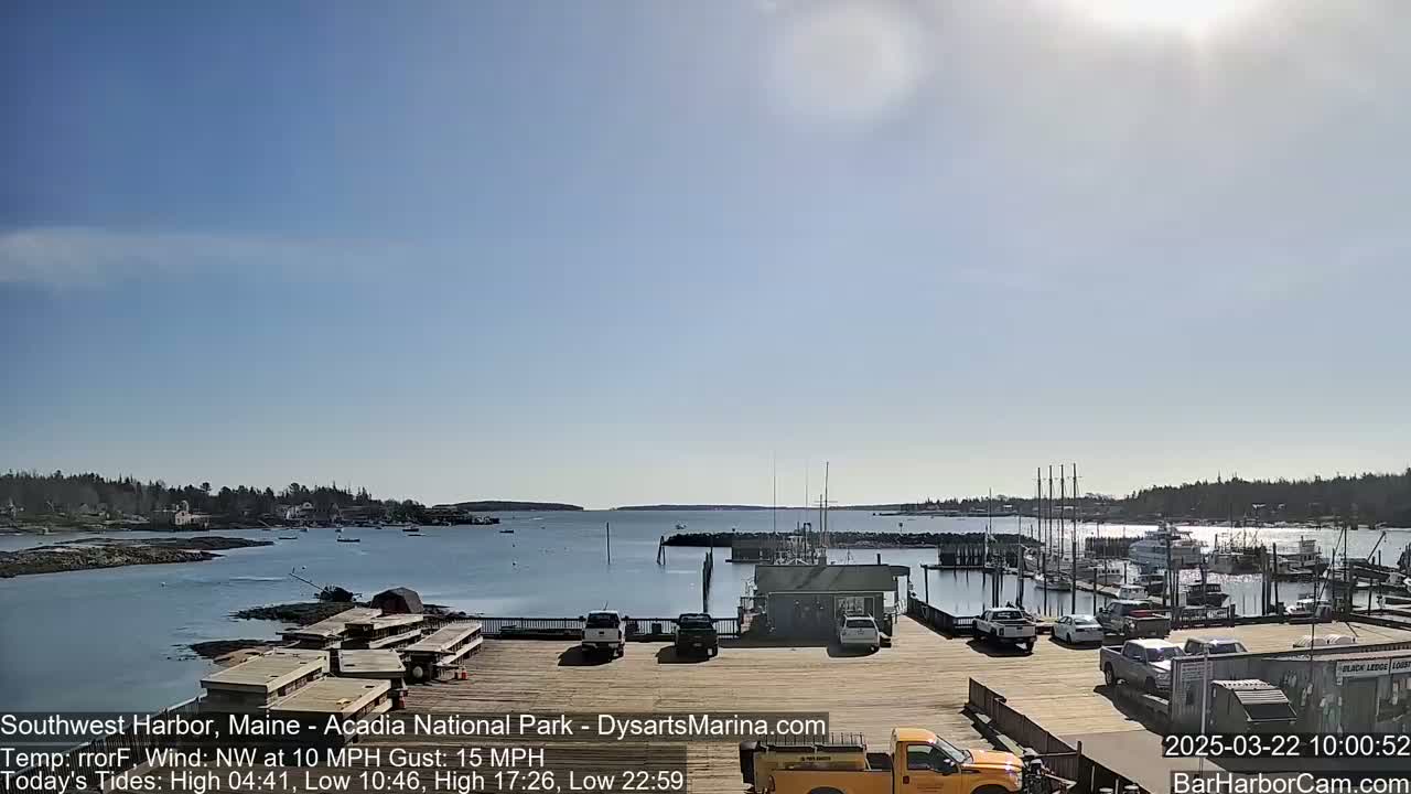 A sunny harbor scene with a calm body of water, boats docked at a marina, and a few vehicles parked on a wooden pier.