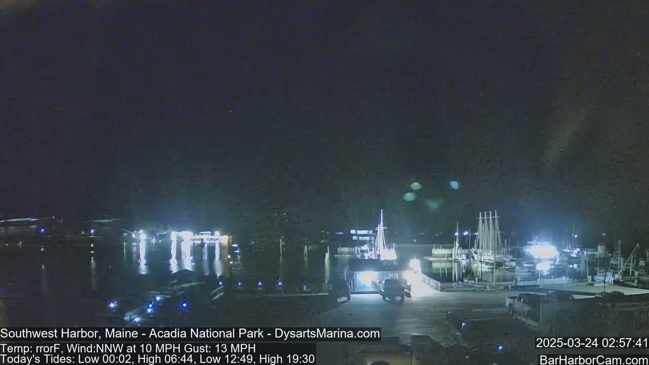 A nighttime harbor scene with numerous boats docked, illuminated by lights under a dark, clear sky with a gentle northwest wind.