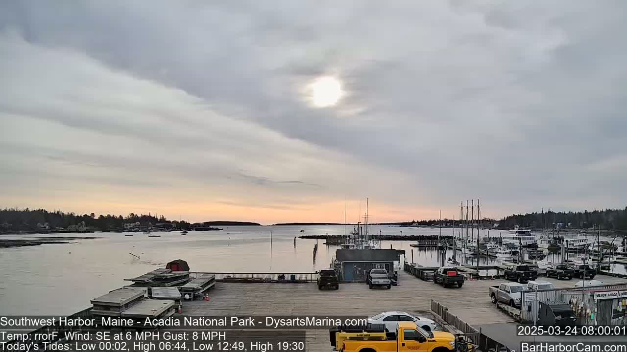 A calm harbor with docked boats is viewed under a mostly cloudy sky with a bright sun, as the sun rises slightly over the horizon.