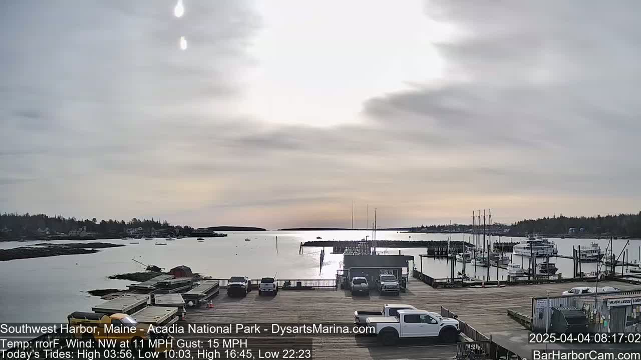A mostly cloudy, calm day overlooks a harbor with docked boats and cars parked in a lot, the sun reflecting on the water.