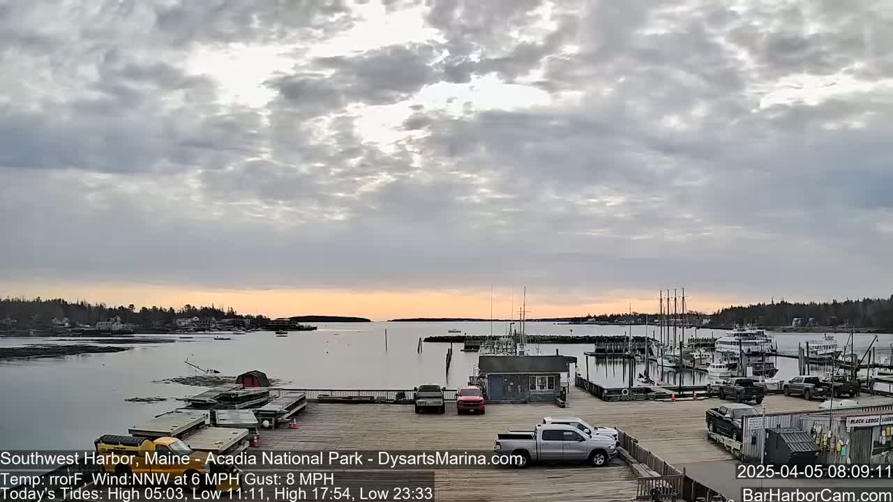 A calm harbor with docked boats is seen at sunrise under a mostly cloudy sky, with a light wind.