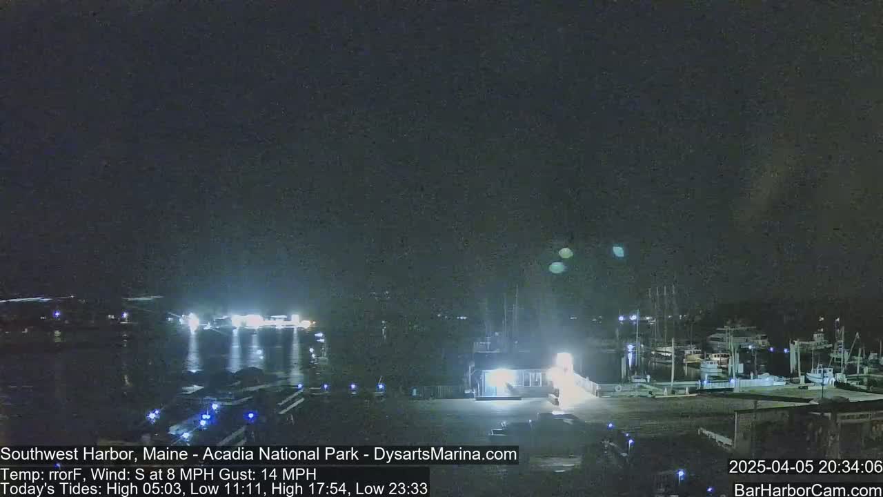 A nighttime view of a harbor with boats docked, illuminated by lights, under a dark sky with a slight south wind.