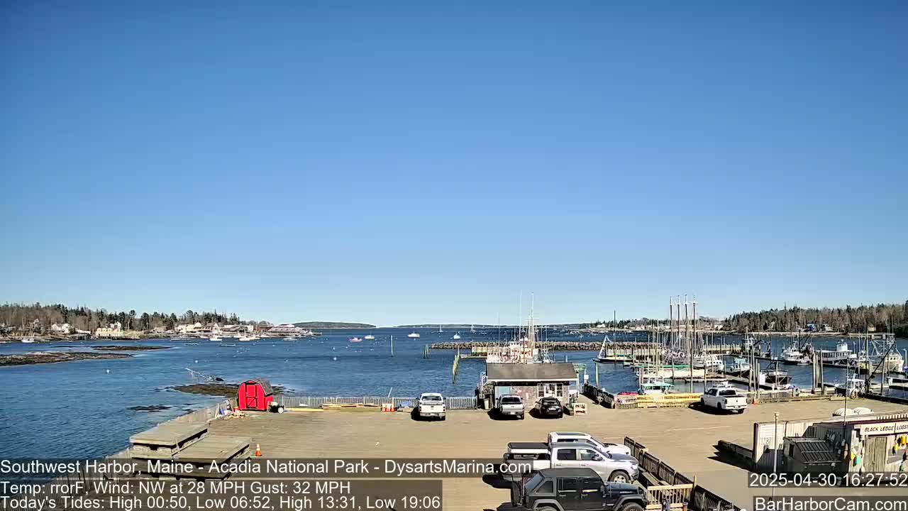 A harbor with boats docked, cars in a parking lot, and a clear blue sky under a bright sun.
