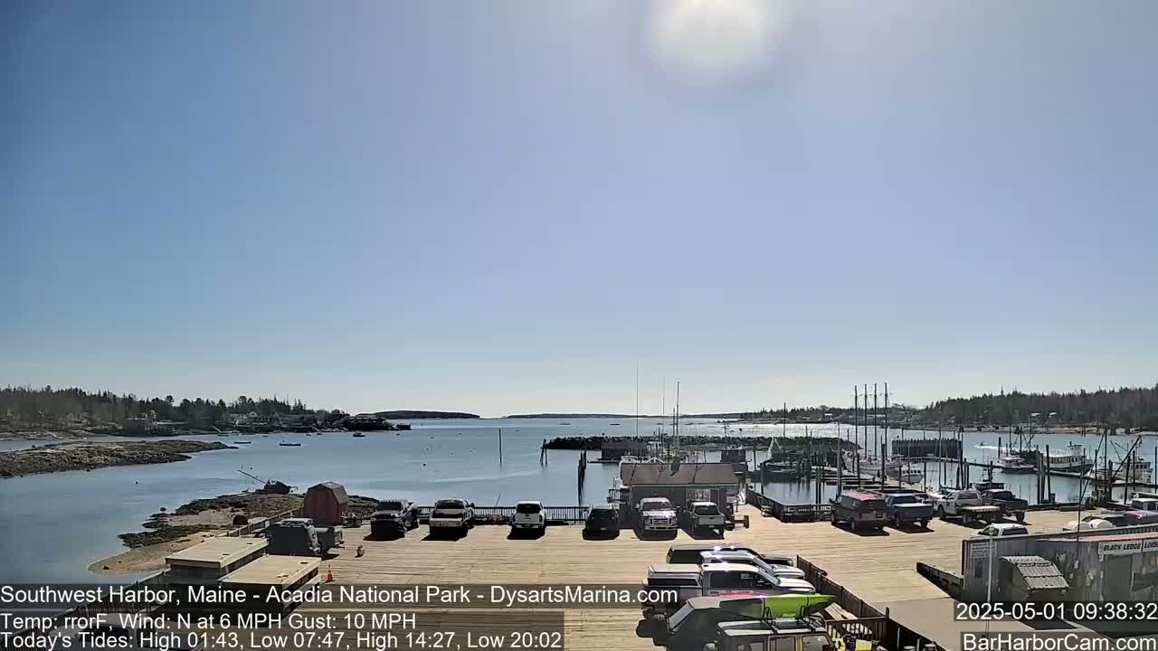 A sunny harbor scene with a calm body of water, boats docked at a marina, and cars parked in a lot, under a clear blue sky.