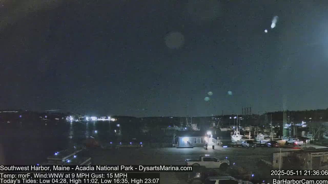 A nighttime harbor scene with several boats docked, under a dark sky with a few bright lights visible in the distance.