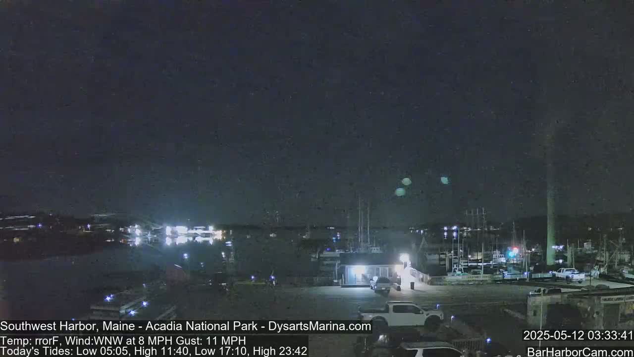 A nighttime view of a harbor with boats docked, a parking lot with vehicles, and a dark sky with a few bright lights.