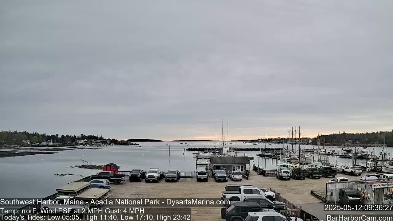 A calm harbor with numerous boats docked, a parking lot filled with cars, and a partly cloudy sky.