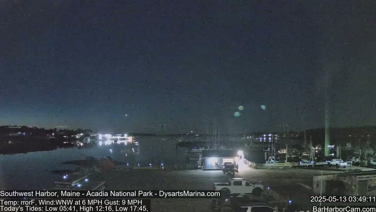 A nighttime view of a harbor with boats and a building, under a mostly clear sky with a few faint lights visible in the distance.