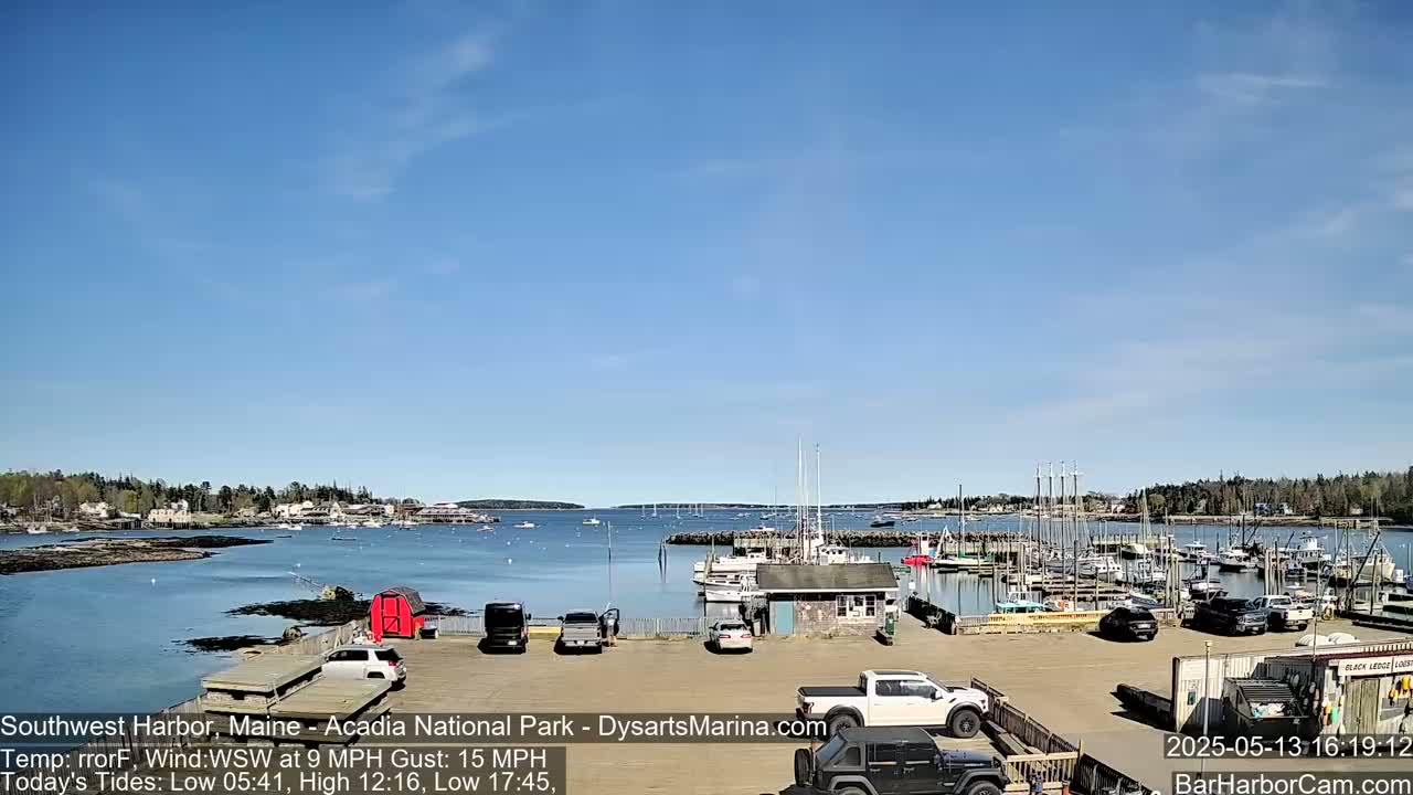 A sunny day reveals a harbor filled with boats and cars parked in a lot, overlooking a calm body of water and a town in the distance.