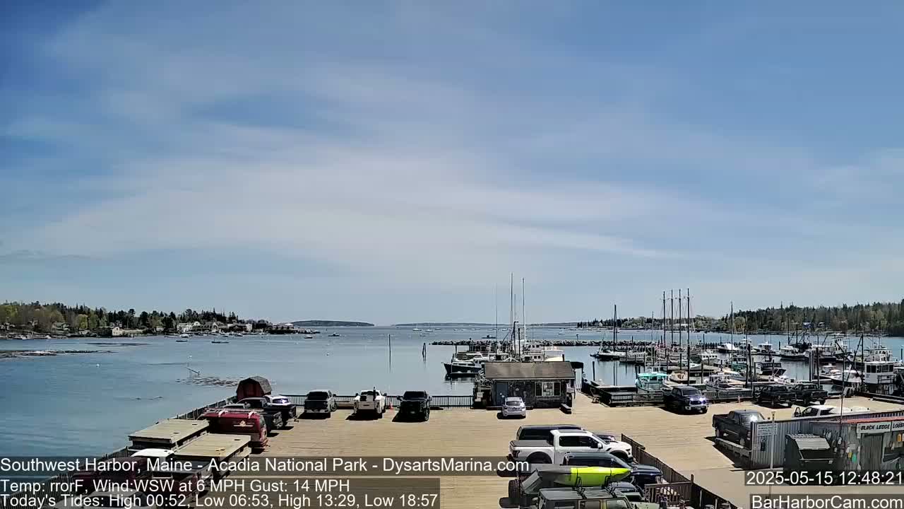 A mostly sunny day shows a harbor filled with docked boats and cars parked in a lot next to the water.