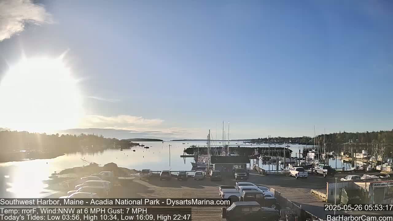 A mostly sunny day shows a harbor filled with boats and cars parked in a lot beside it.