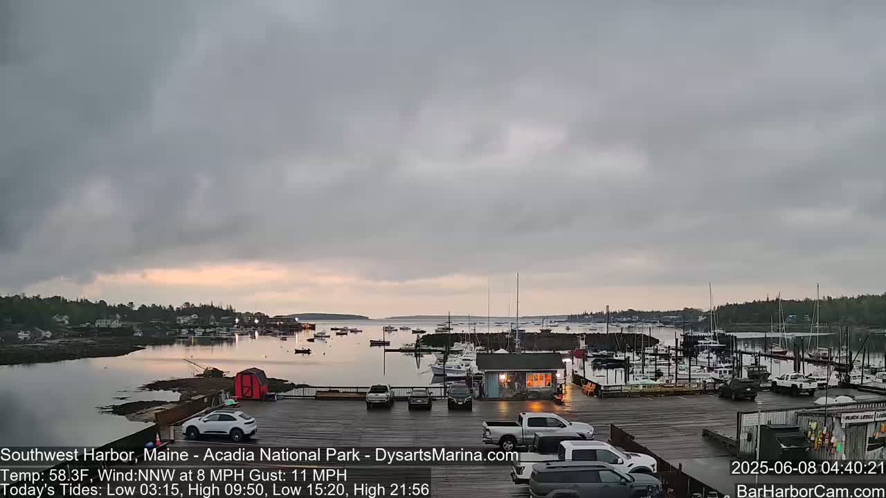 A mostly cloudy sunrise over a harbor filled with boats, with cars parked in a lot next to a small building.