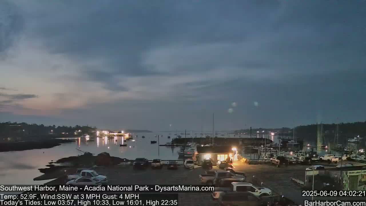A twilight view of a harbor filled with boats, with cars parked in a lot nearby under a mostly cloudy sky.